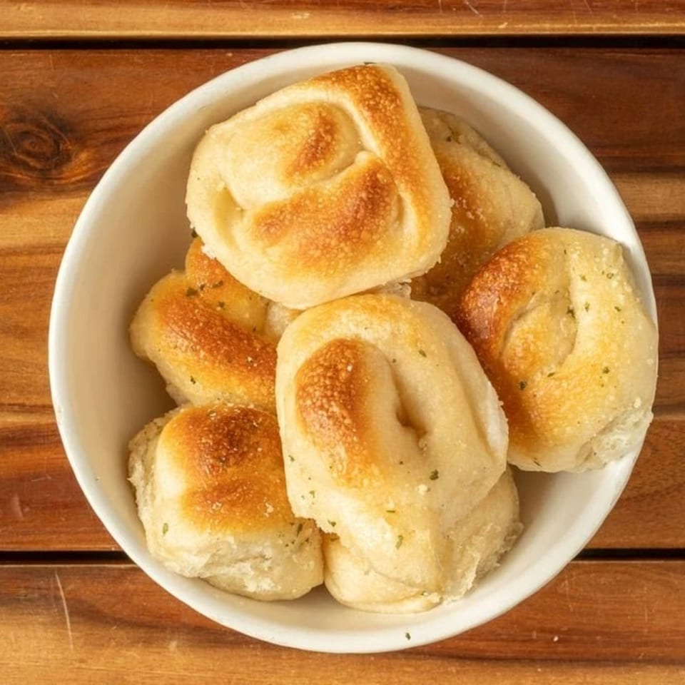 A white bowl filled with golden-brown garlic knots, sprinkled with herbs, on a wooden table.
