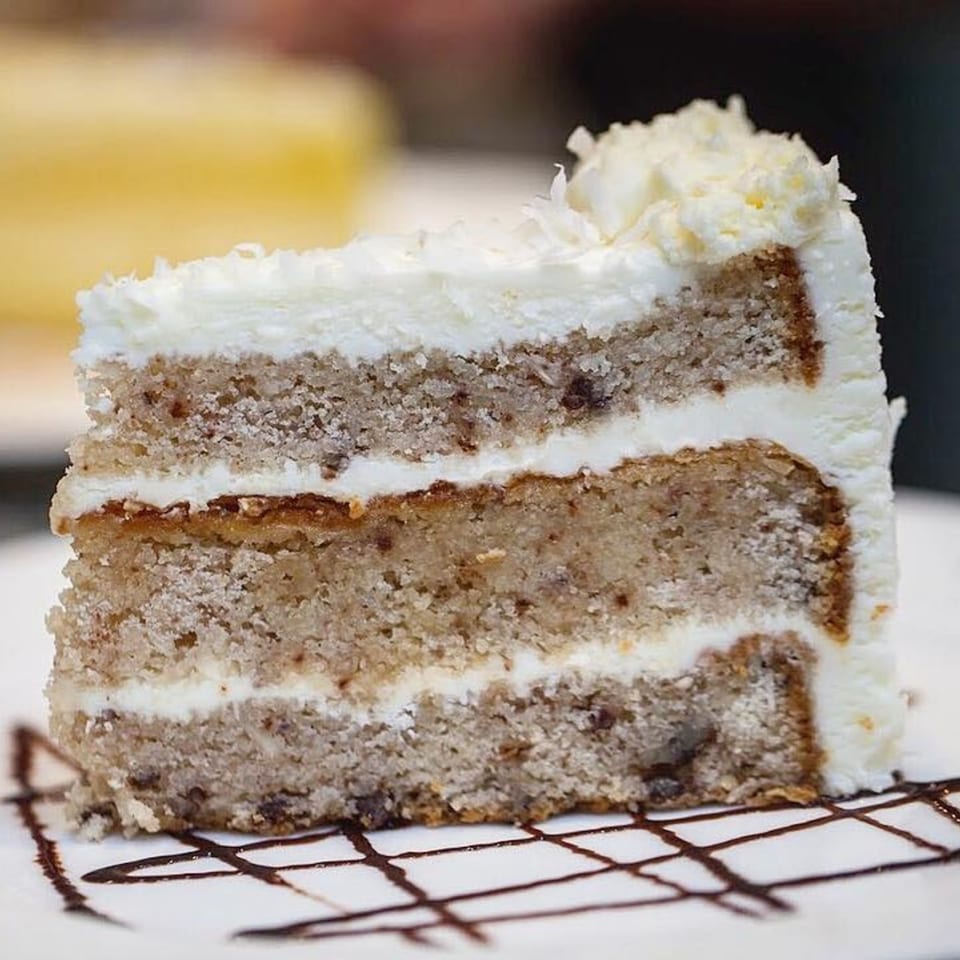 A slice of layered cake with white frosting and chocolate drizzle on a white plate.