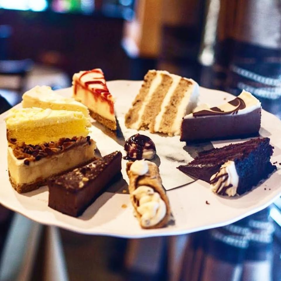 A white plate holds various dessert slices, including cheesecake, chocolate cake, and cannoli.