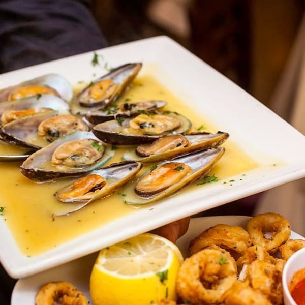 Steamed mussels in a white wine sauce on a rectangular plate, with fried calamari and lemon below.