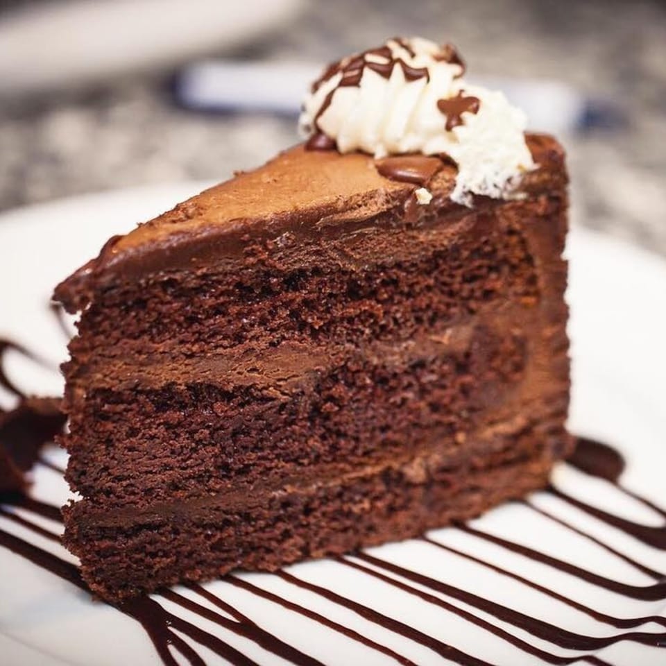 A slice of rich chocolate cake with chocolate frosting, whipped cream, and chocolate drizzle on a white plate.