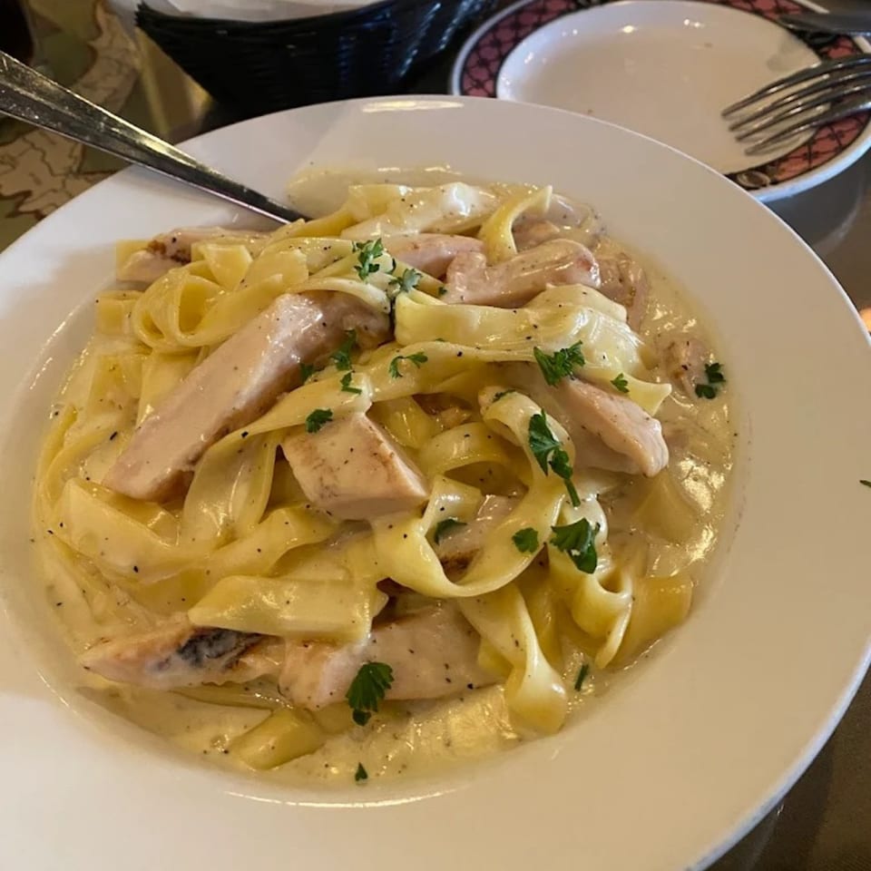A white bowl of creamy fettuccine alfredo with chicken and parsley, a fork on the side.